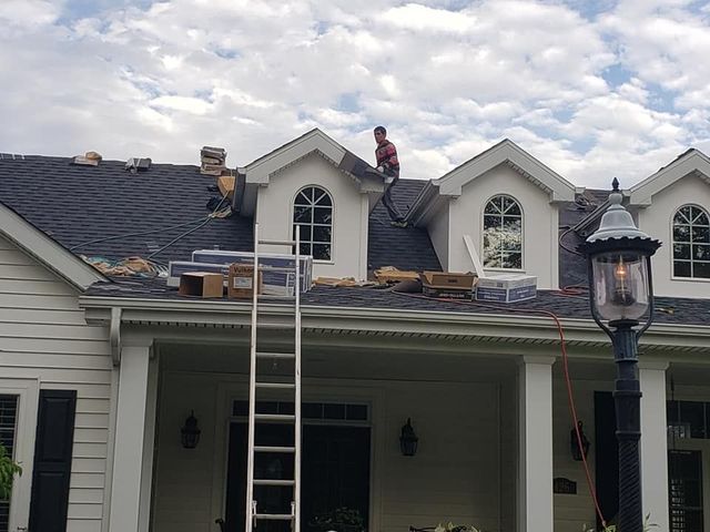 A worker repairs the roof of a house with several gabled dormers on a bright, cloudy day.