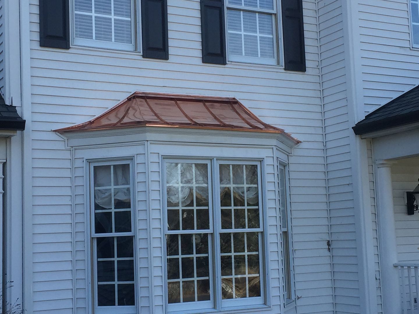A white house exterior featuring a copper-roofed bay window topped by two rectangular windows with black shutters.