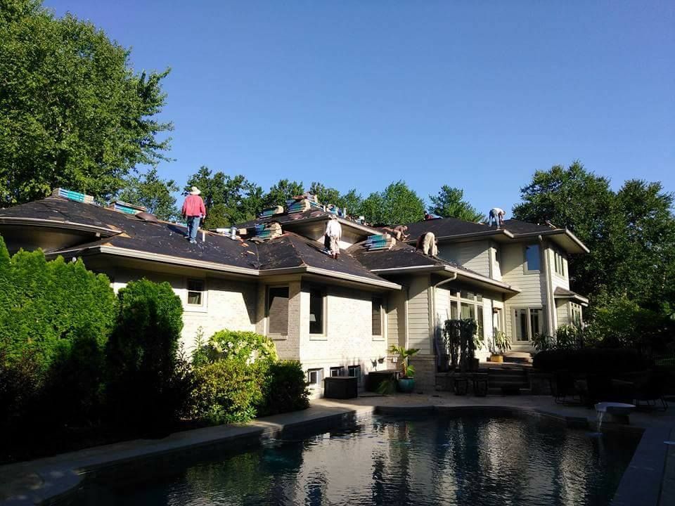 Workers install new shingles on the roof of a large, light-colored house next to a backyard swimming pool.