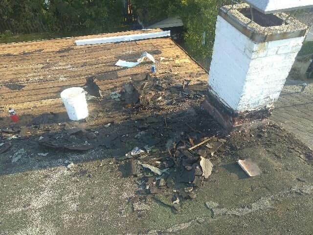 A damaged rooftop with a white chimney, scattered roofing debris, and a white bucket on the exposed wooden sheathing.