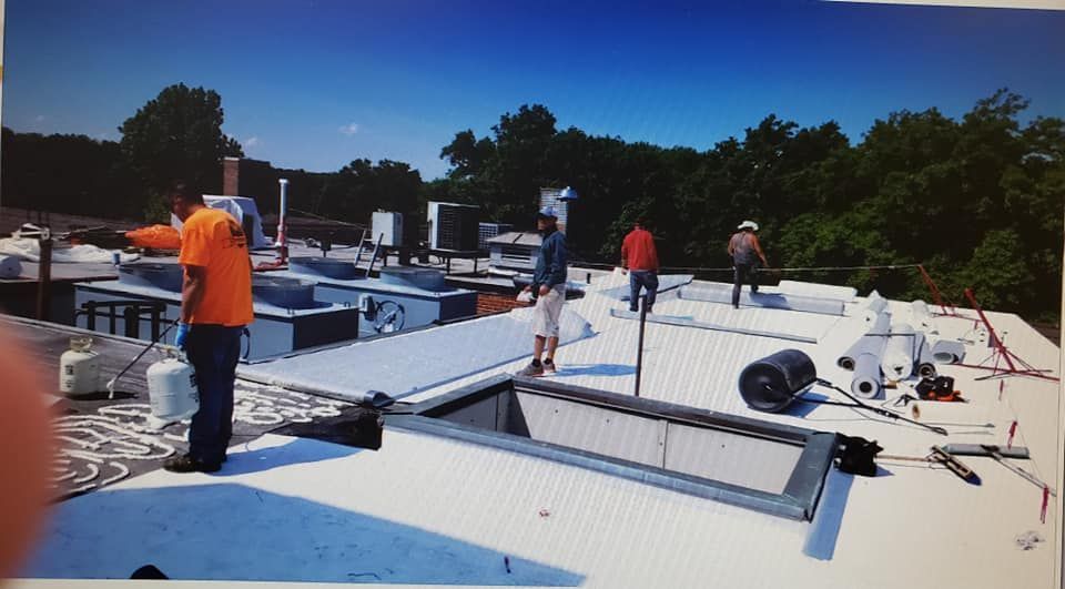 Four workers in high-visibility orange shirts install or repair a flat, white commercial roof on a sunny day.