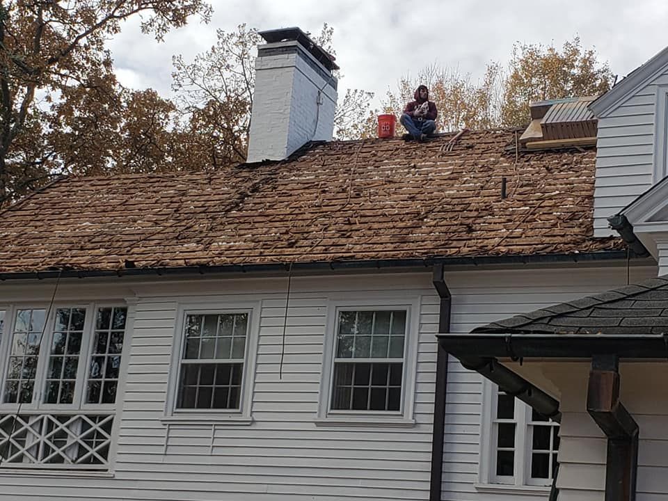 A person sits on a wooden roof near a brick chimney on a white, two-story house surrounded by trees.