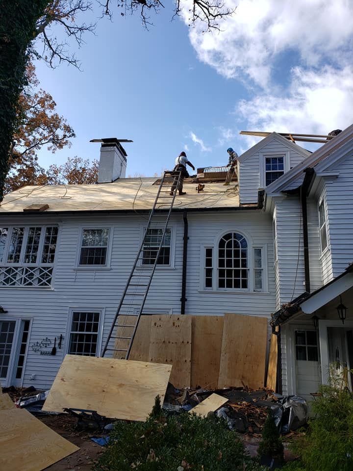 Two workers on a ladder and the roof of a large white house during a roof renovation project.