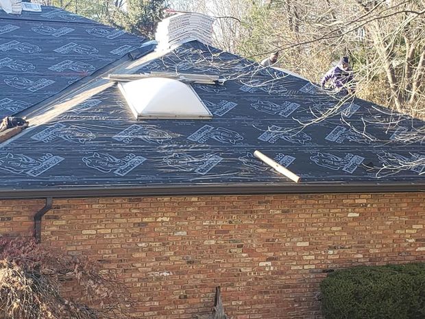 Workers installing black underlayment on a residential roof featuring a chimney and a white skylight.