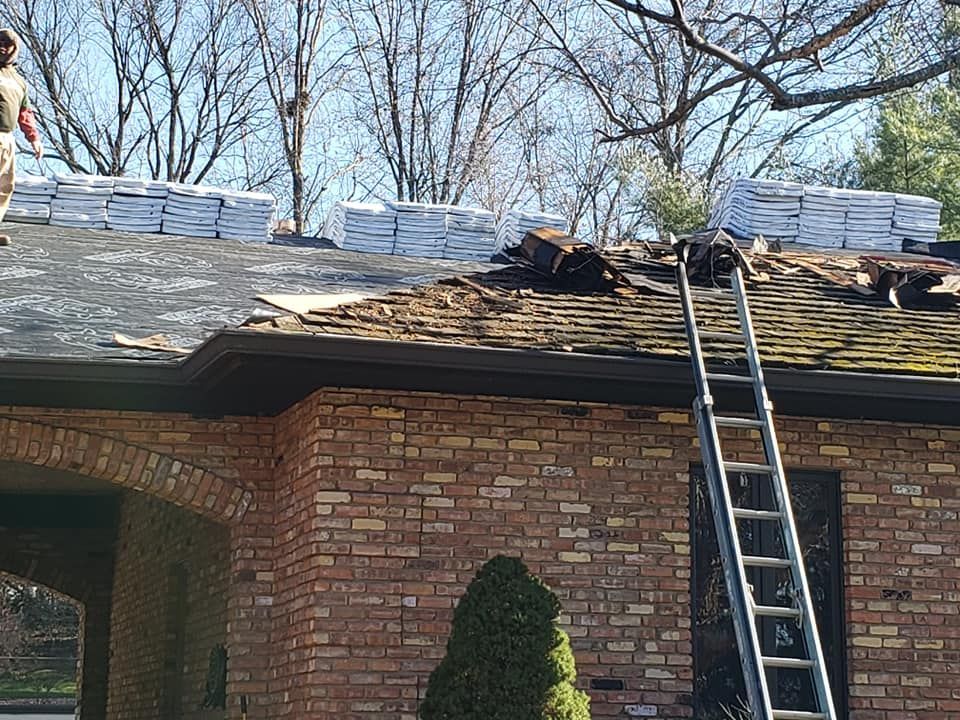A roof replacement project in progress with stacks of shingles, a ladder, and bare roof patches on a brick house.