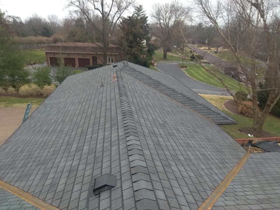 An elevated view of a gray asphalt shingled roof with a central ridge vent, overlooking a driveway and wooded yard.