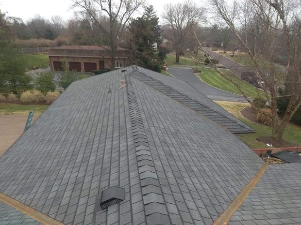 An elevated view of a gray asphalt shingled roof with a central ridge vent, overlooking a driveway and wooded yard.