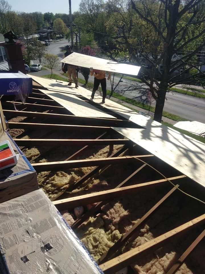 Two people in hard hats install plywood sheathing on a residential roof frame over open rafters.