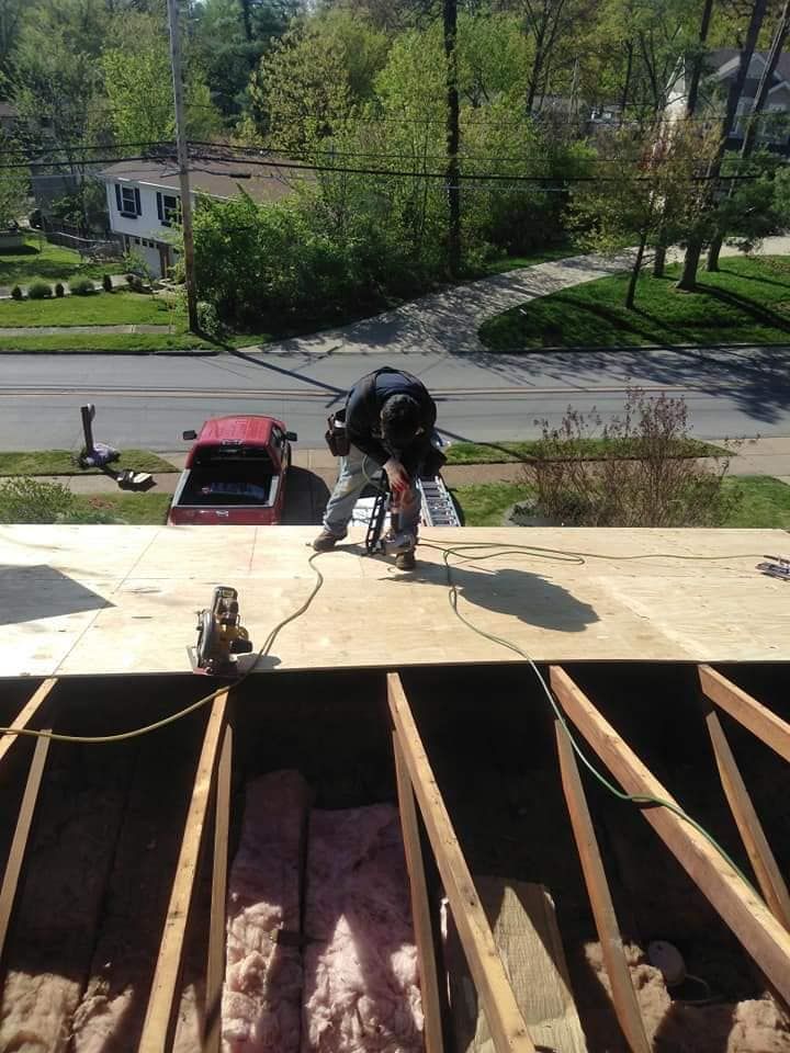 A worker cuts a piece of plywood on a residential roof framework with an open attic visible below.
