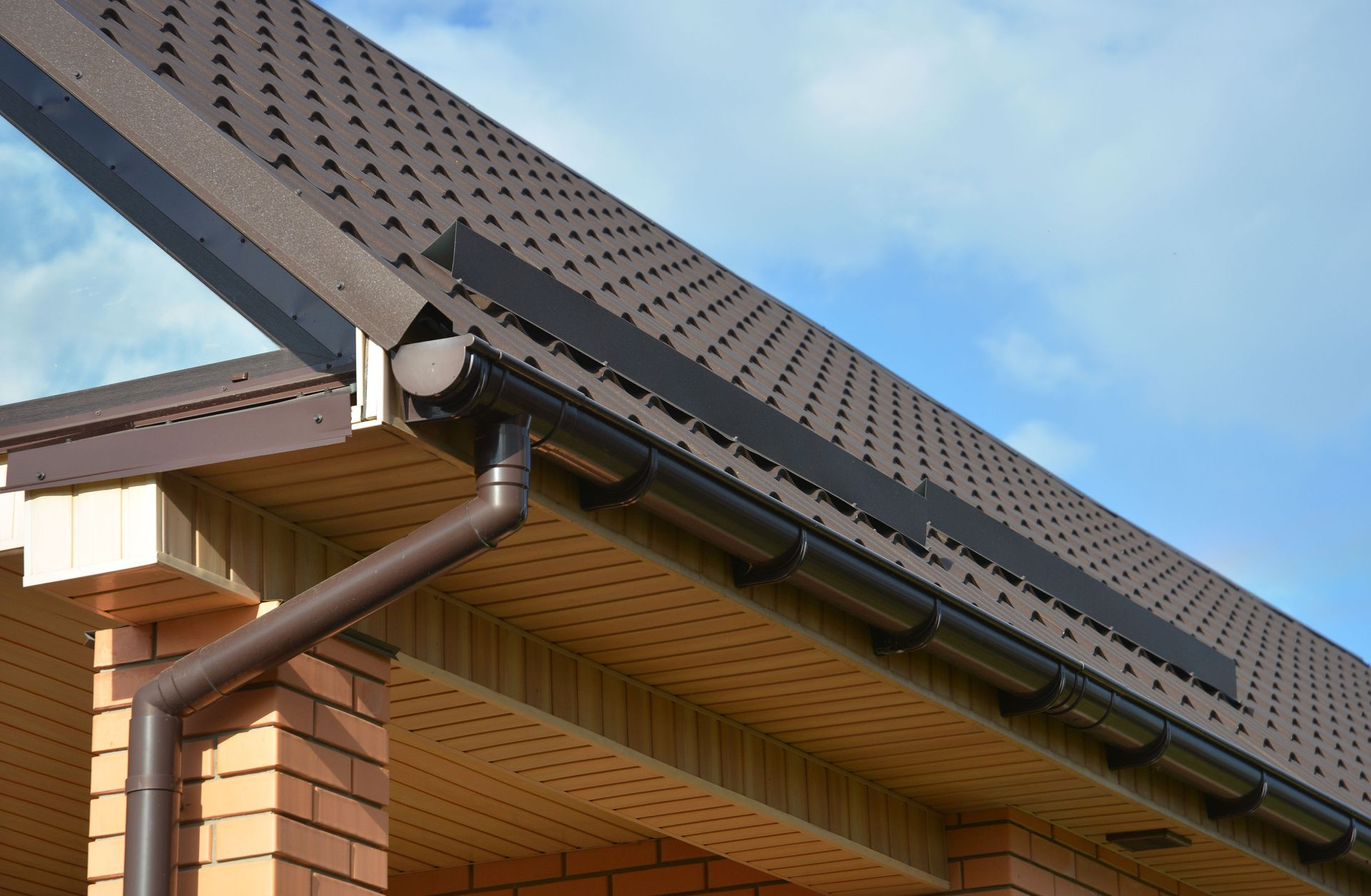 Brown metal roof panels with a matching gutter and downspout system installed on a brick building under a cloudy sky.