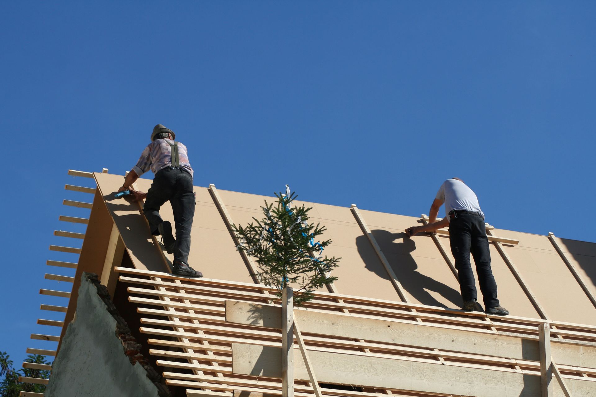 Two people stand on the wooden frame of a roof under construction against a clear blue sky.