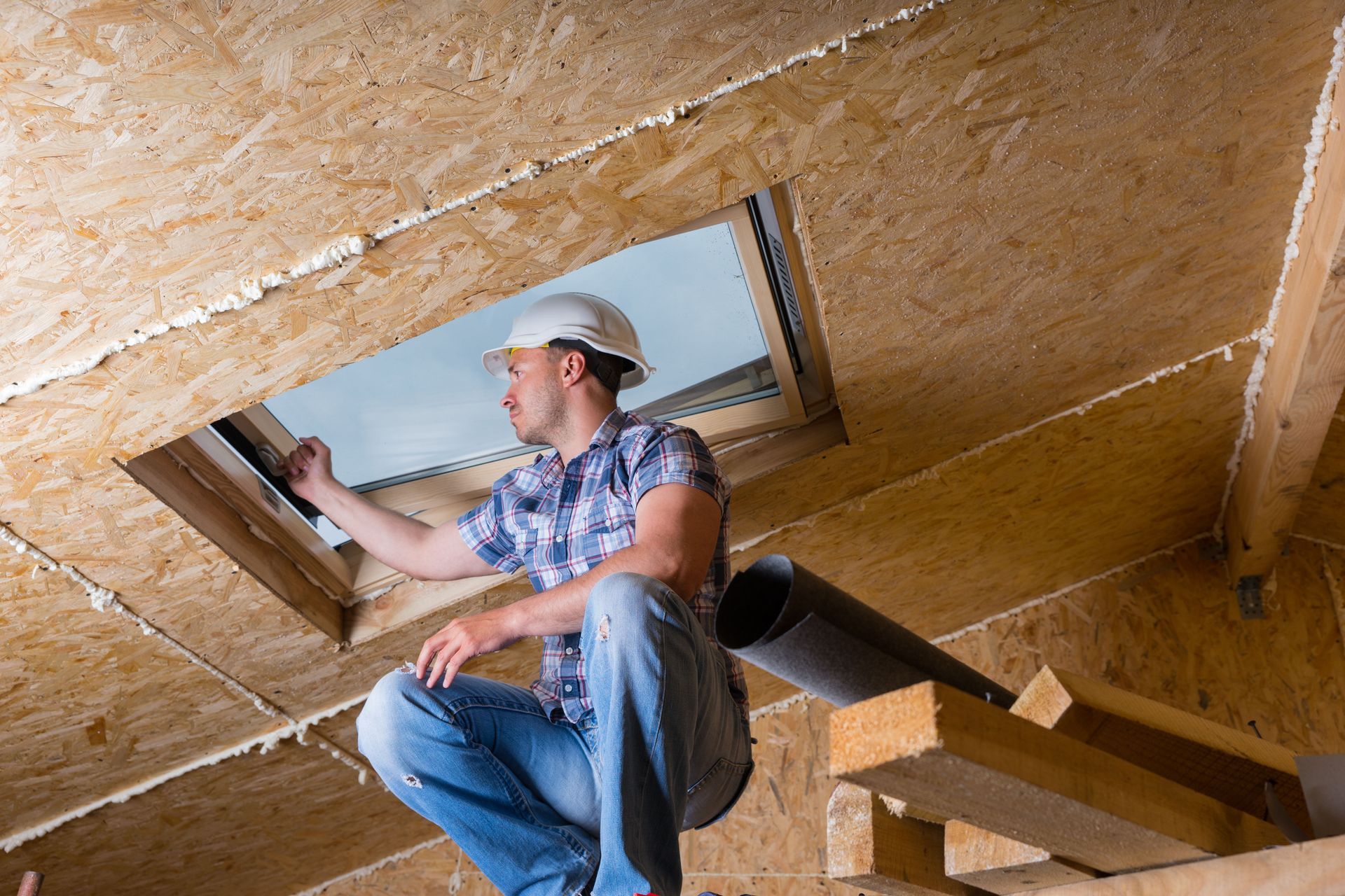 A worker in a hard hat inspects a skylight in an unfinished wooden attic space.