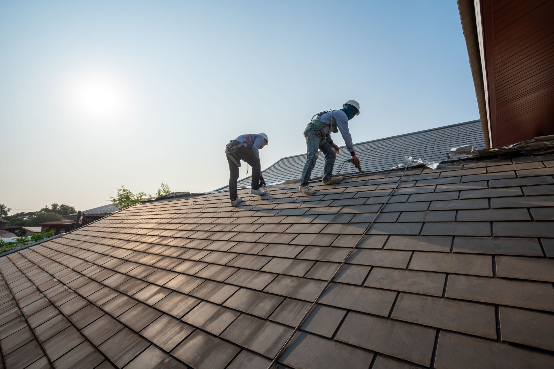 Two construction workers in safety gear install tiles on a residential roof during a sunny day.