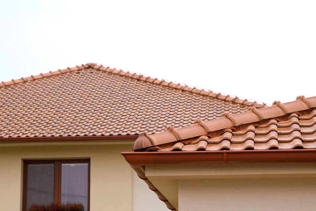 A partial view of two beige houses with terracotta roof tiles and a brown gutter under a plain white sky.