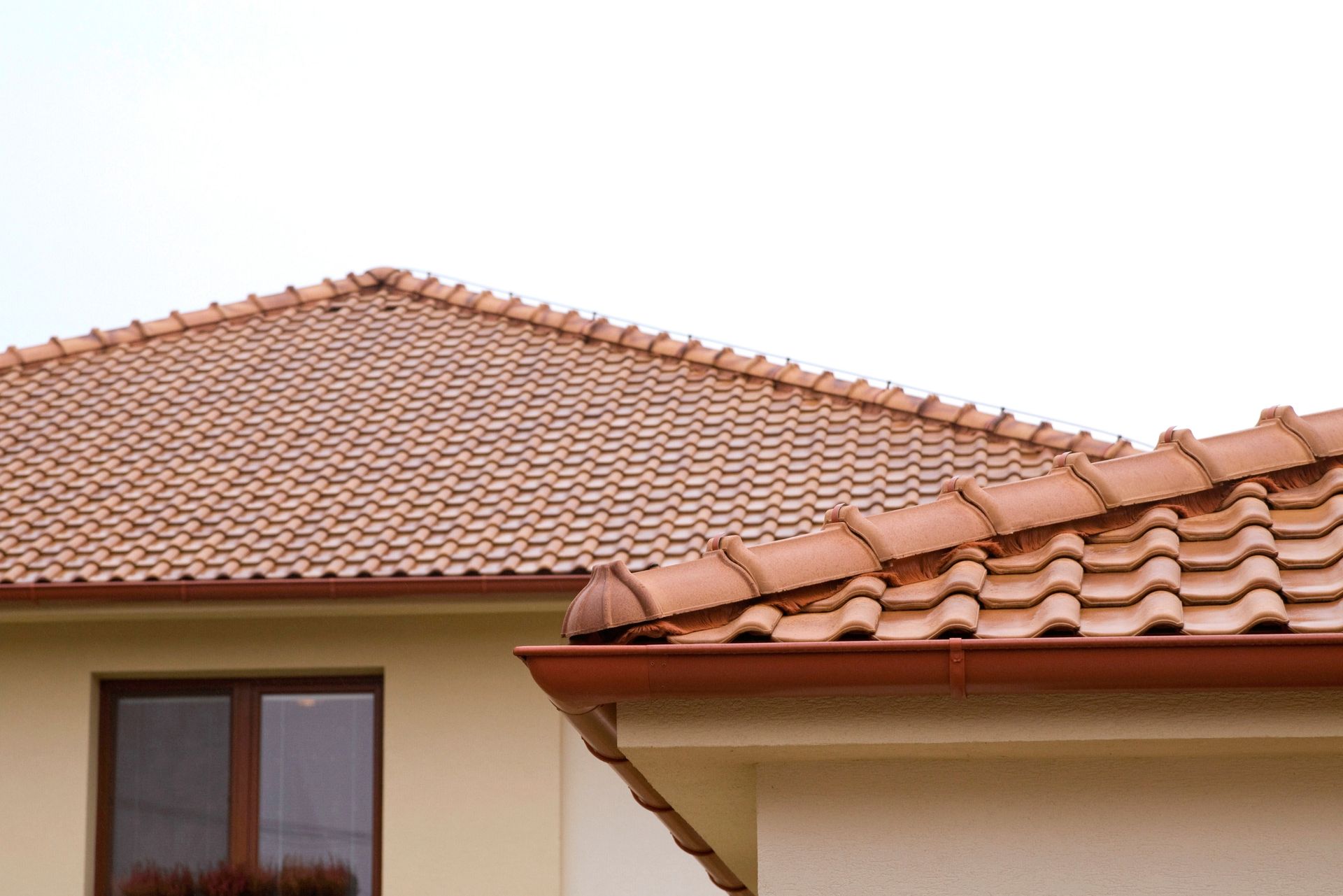 A partial view of two beige houses with terracotta roof tiles and a brown gutter under a plain white sky.