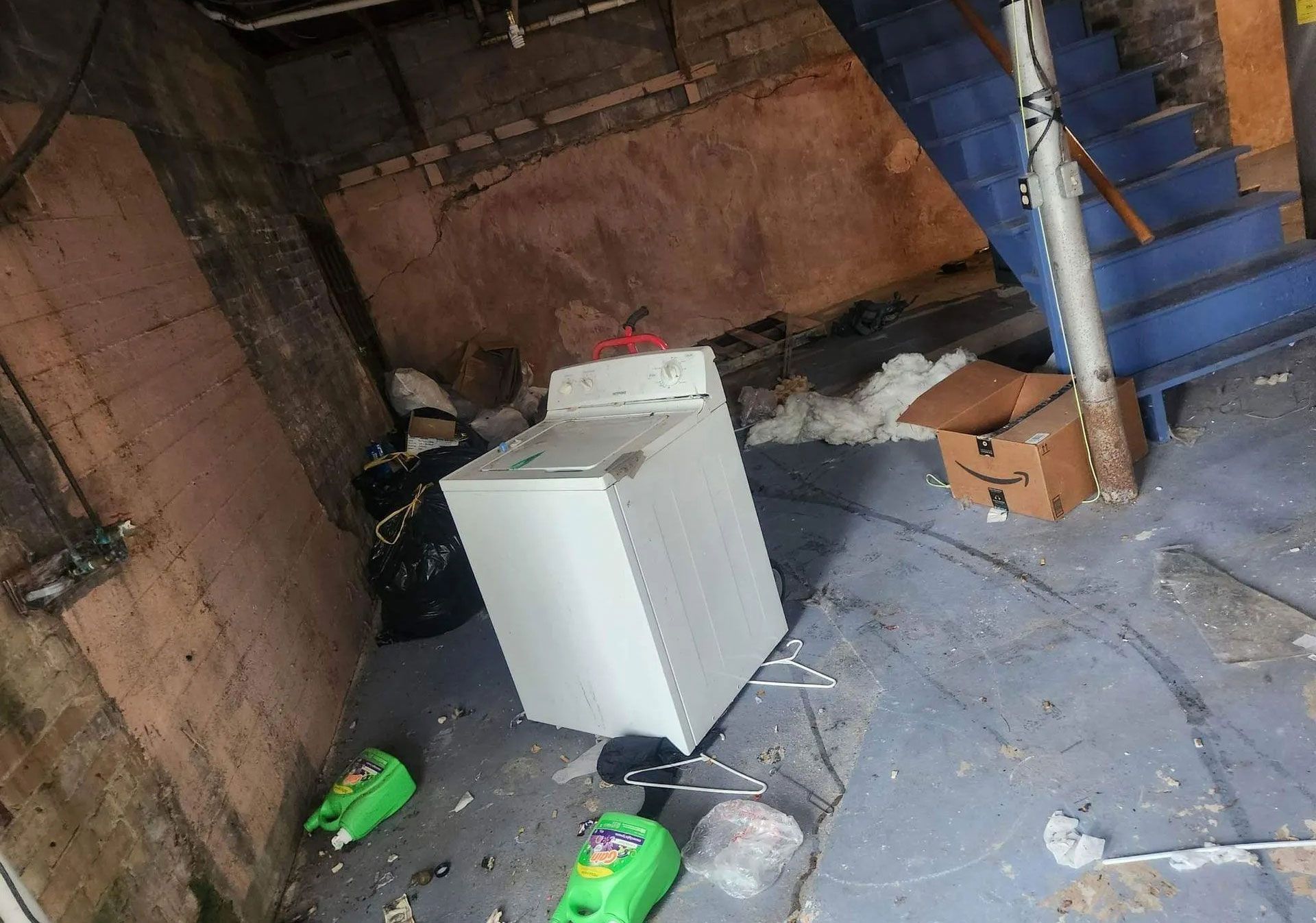 A white washing machine stands in a cluttered basement with unfinished brick walls, blue stairs, and spilled cleaners.