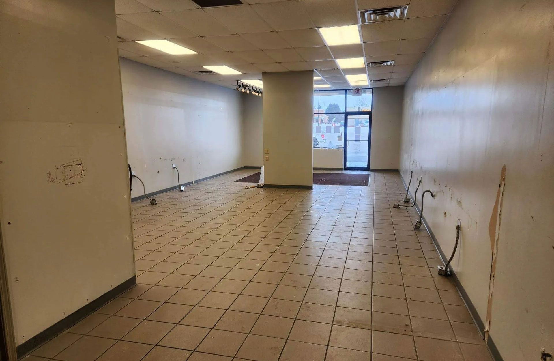 An empty, beige-tiled retail space with white walls, exposed utility pipes, a central support pillar, and glass entryway.