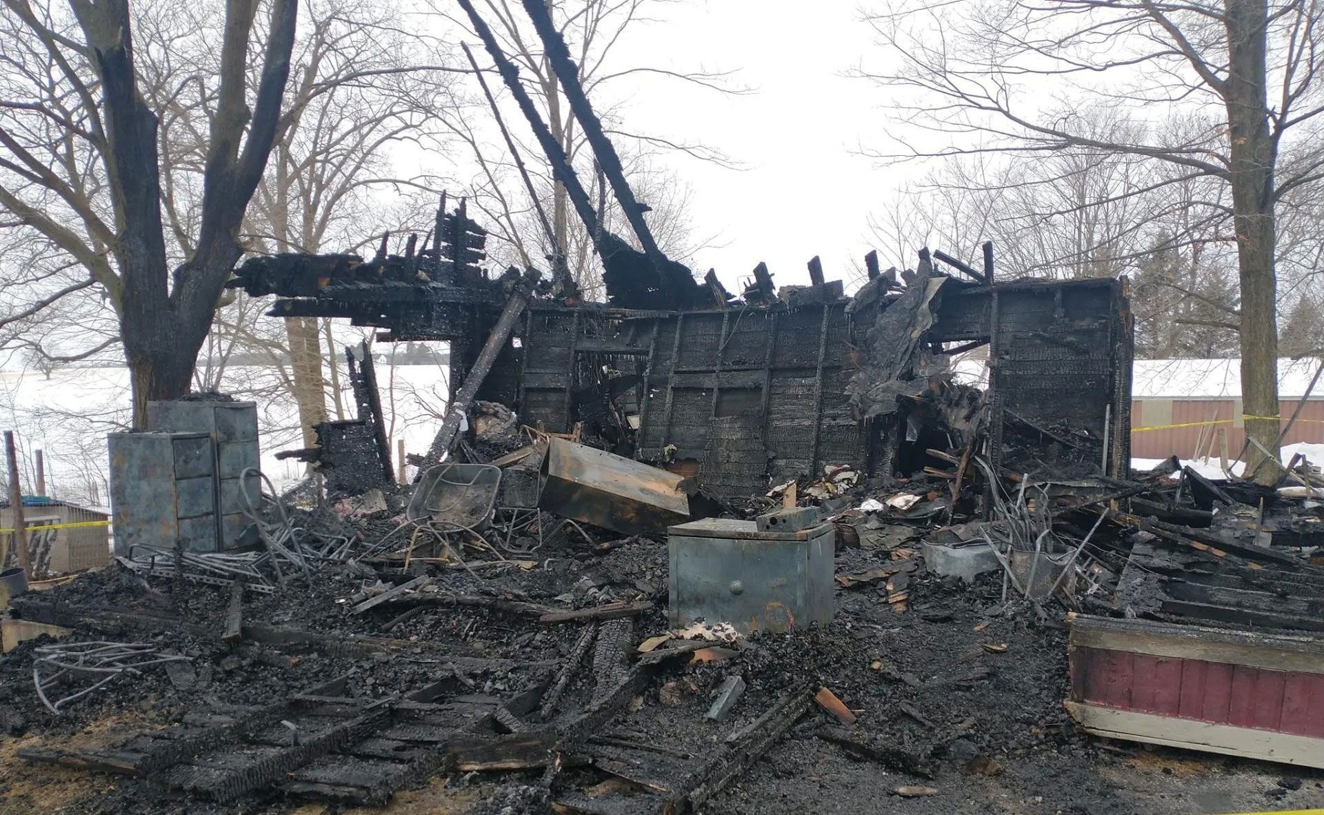 The charred, structural remains of a building destroyed by fire, surrounded by bare trees on a snowy, overcast day.