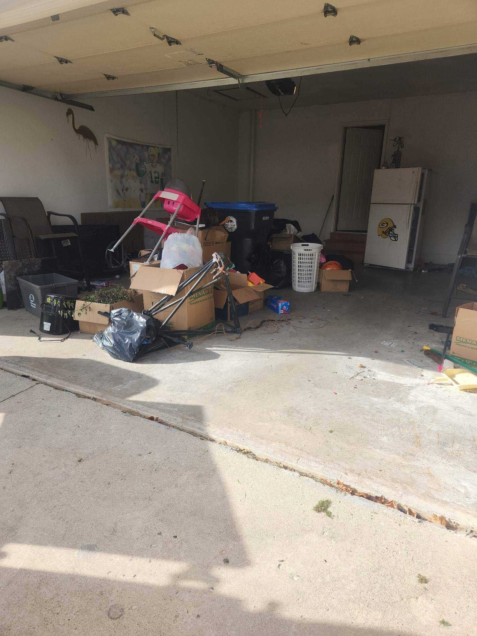 A cluttered residential garage filled with cardboard boxes, a lawn chair, trash cans, a laundry basket, and yard items.