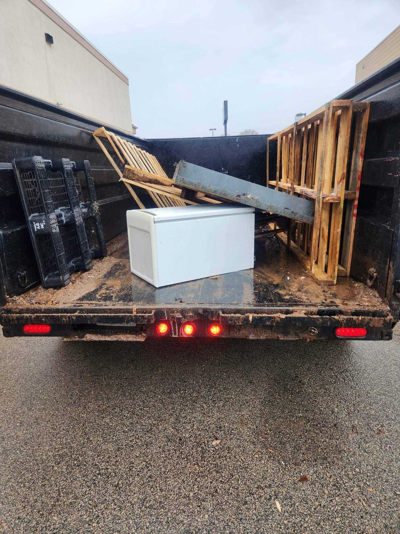 A dump truck bed holding a white chest freezer, a metal beam, and wooden pallets on a wet asphalt surface.