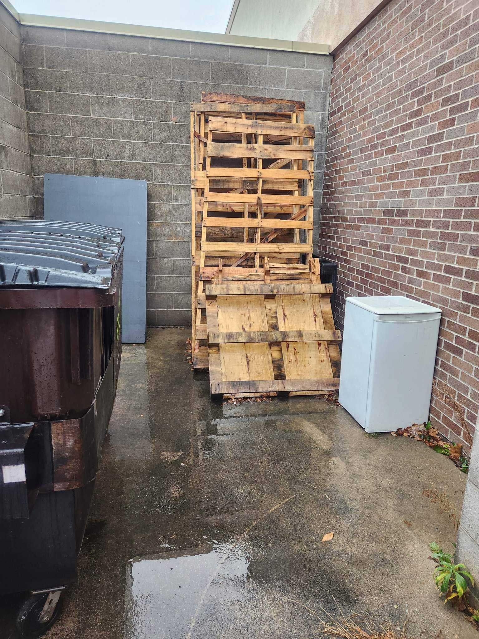 Stacked wooden pallets in an outdoor area between a brick wall, a gray wall, a large dumpster, and a small white cabinet.