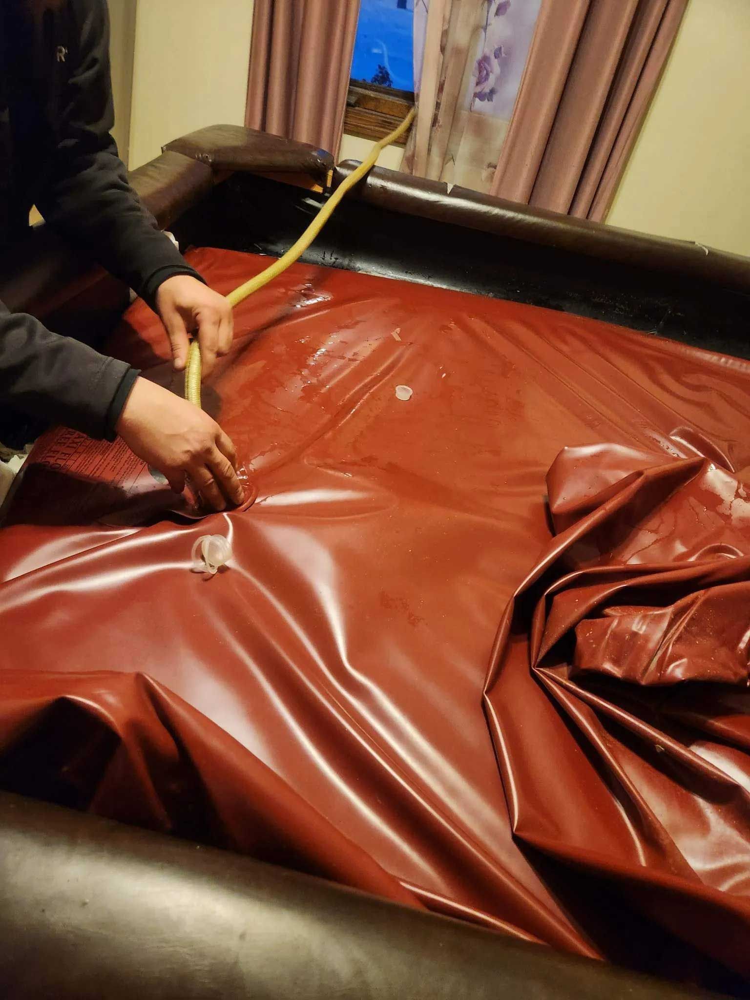 Hands filling a red waterbed mattress with a hose inside a room.