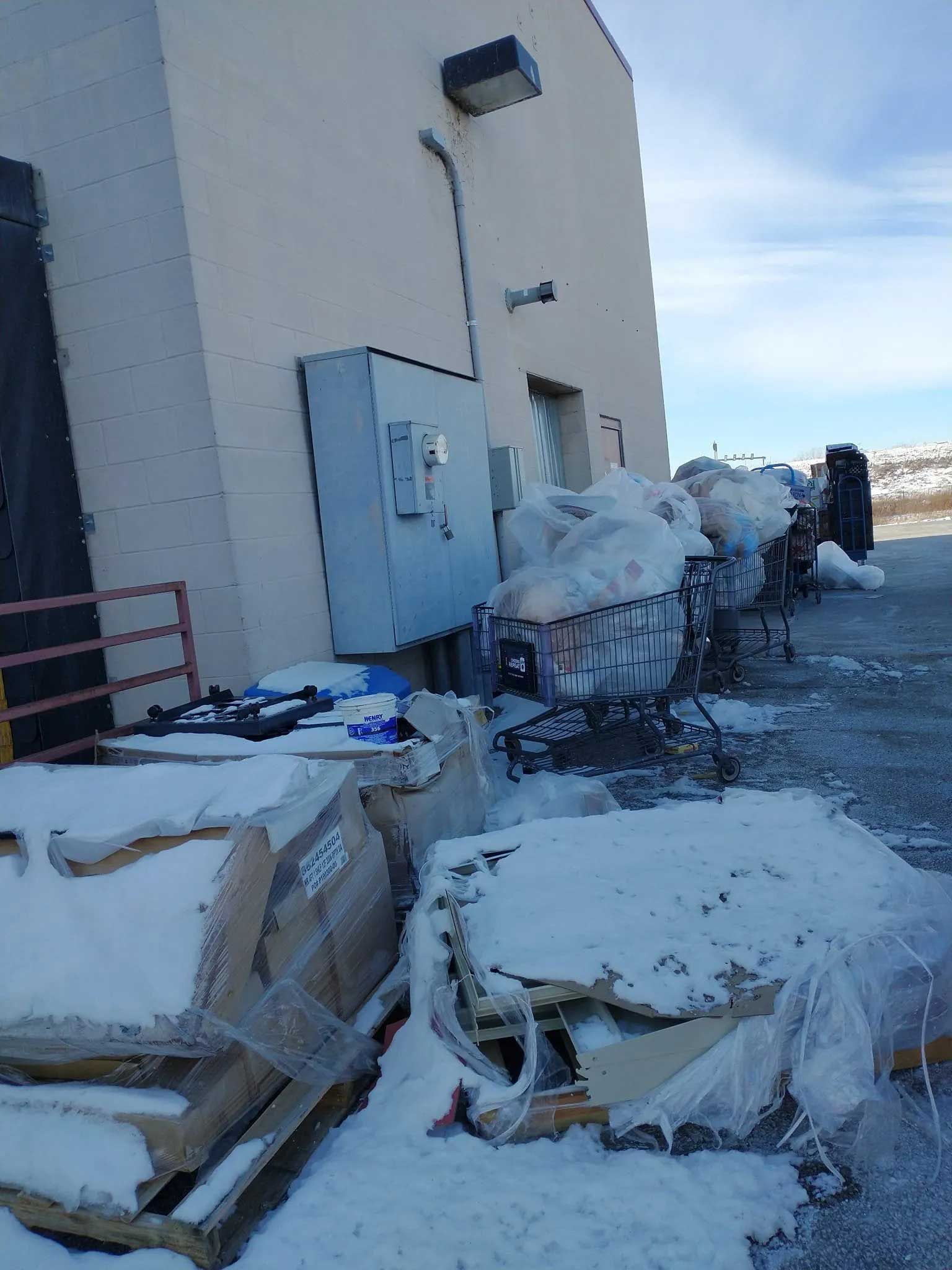 Shopping carts filled with white bags and pallets covered in snow sit outside a beige building.