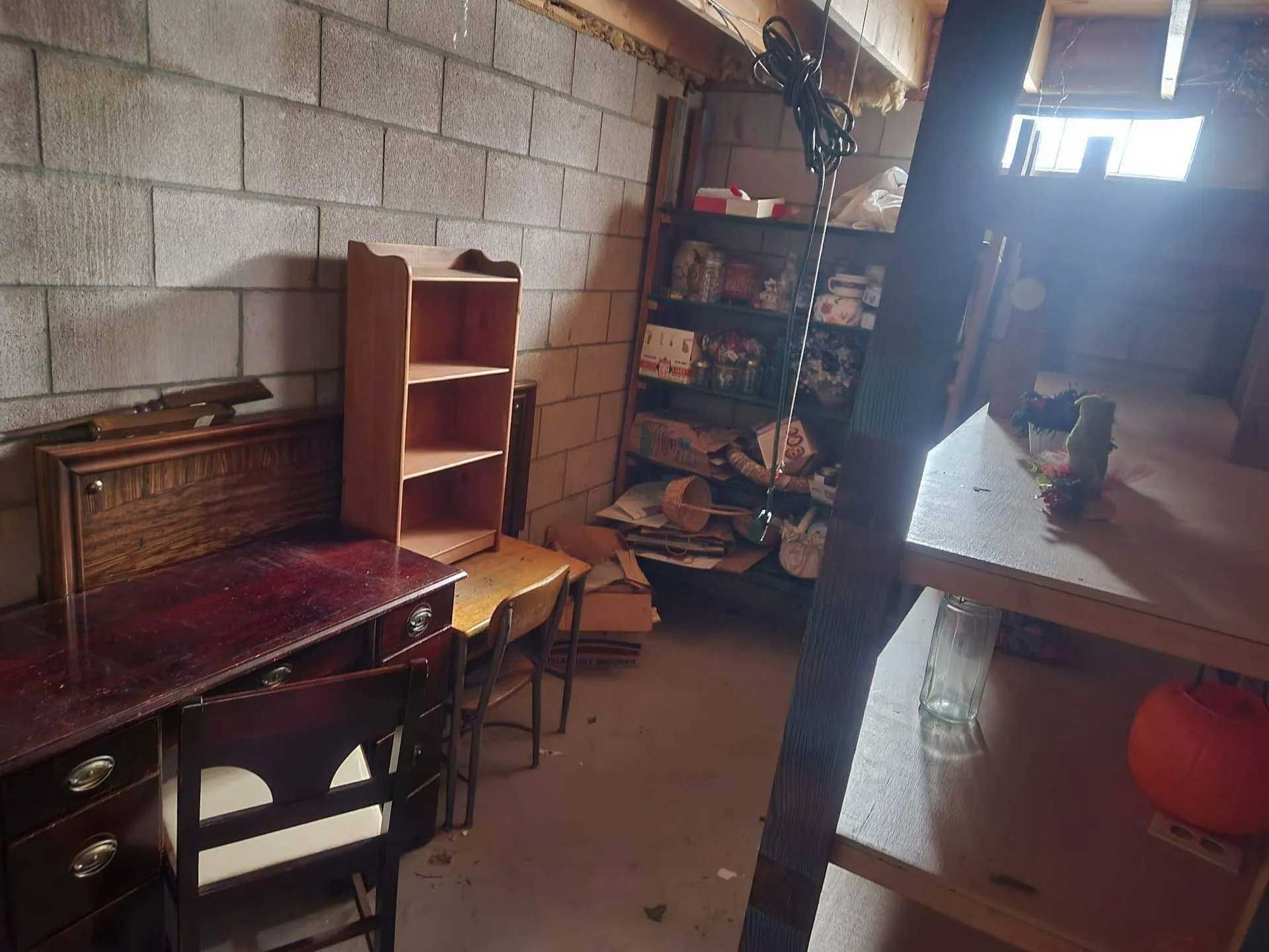 A cluttered storage area with concrete block walls, a desk, a wooden shelving unit, and a metal shelf in the foreground.