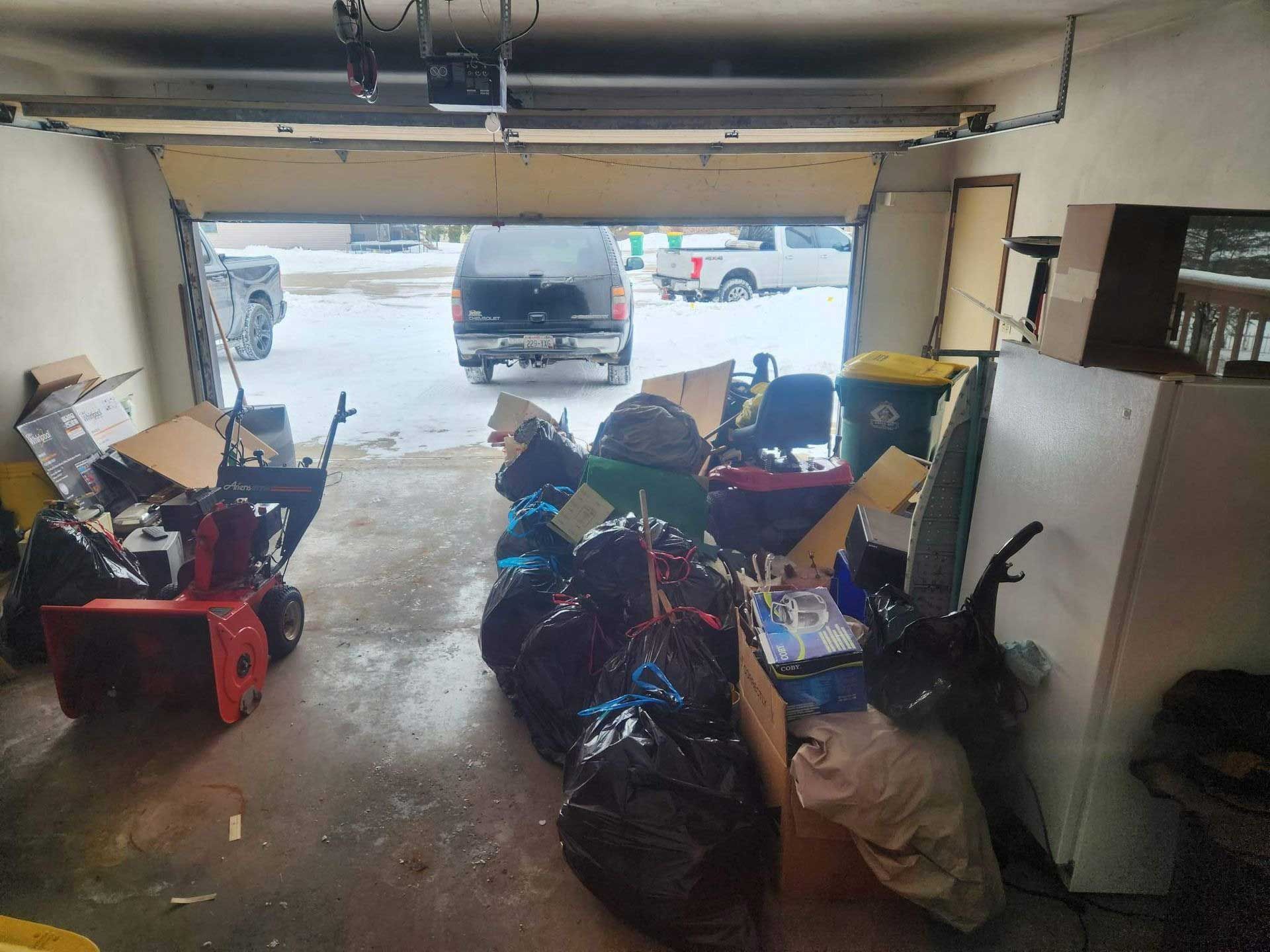 A cluttered garage with a red snowblower on the left, a pile of trash bags in the center, and a white refrigerator.