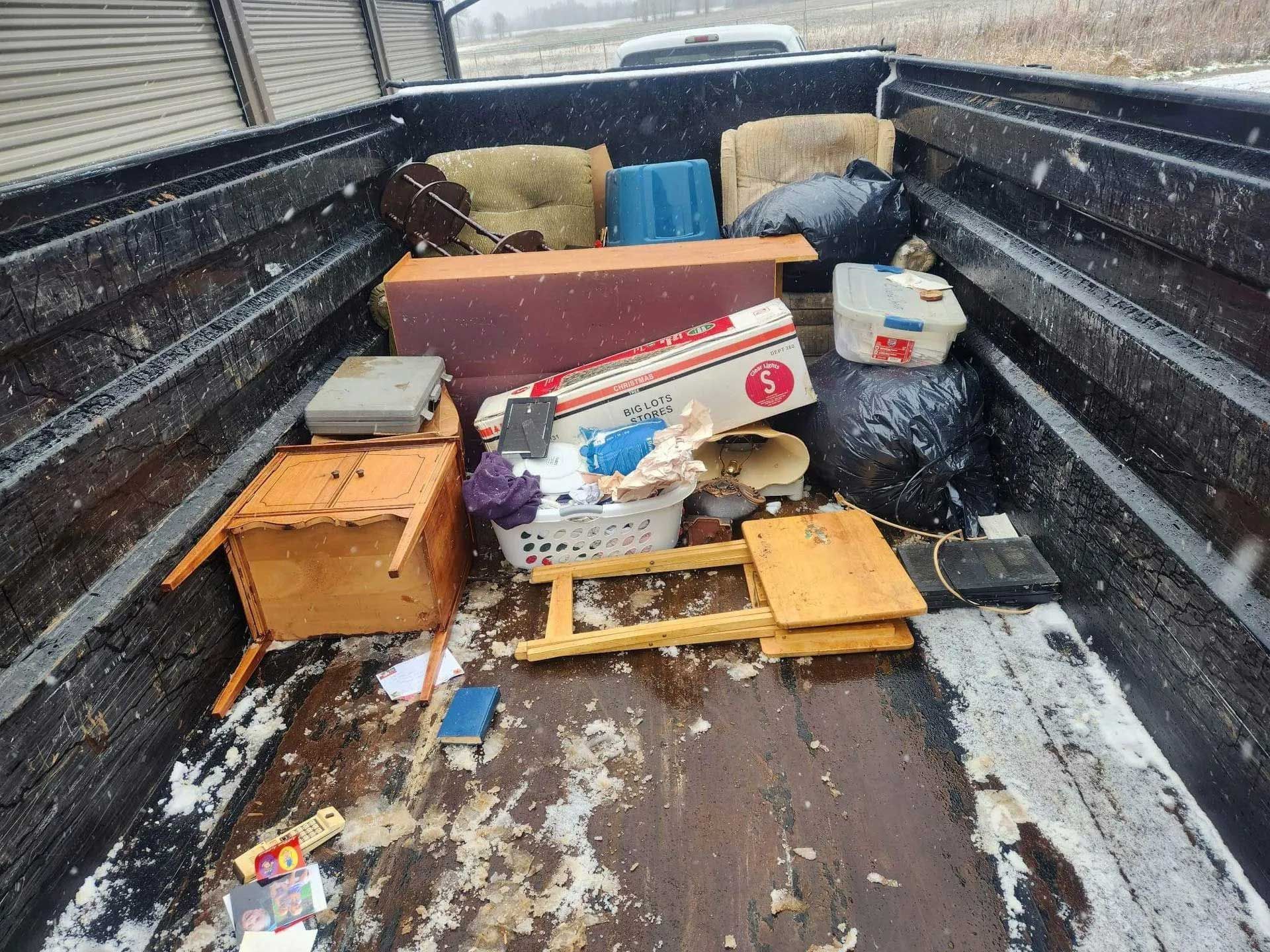 The bed of a black pickup truck loaded with miscellaneous household furniture, plastic bins, and trash bags in the snow.