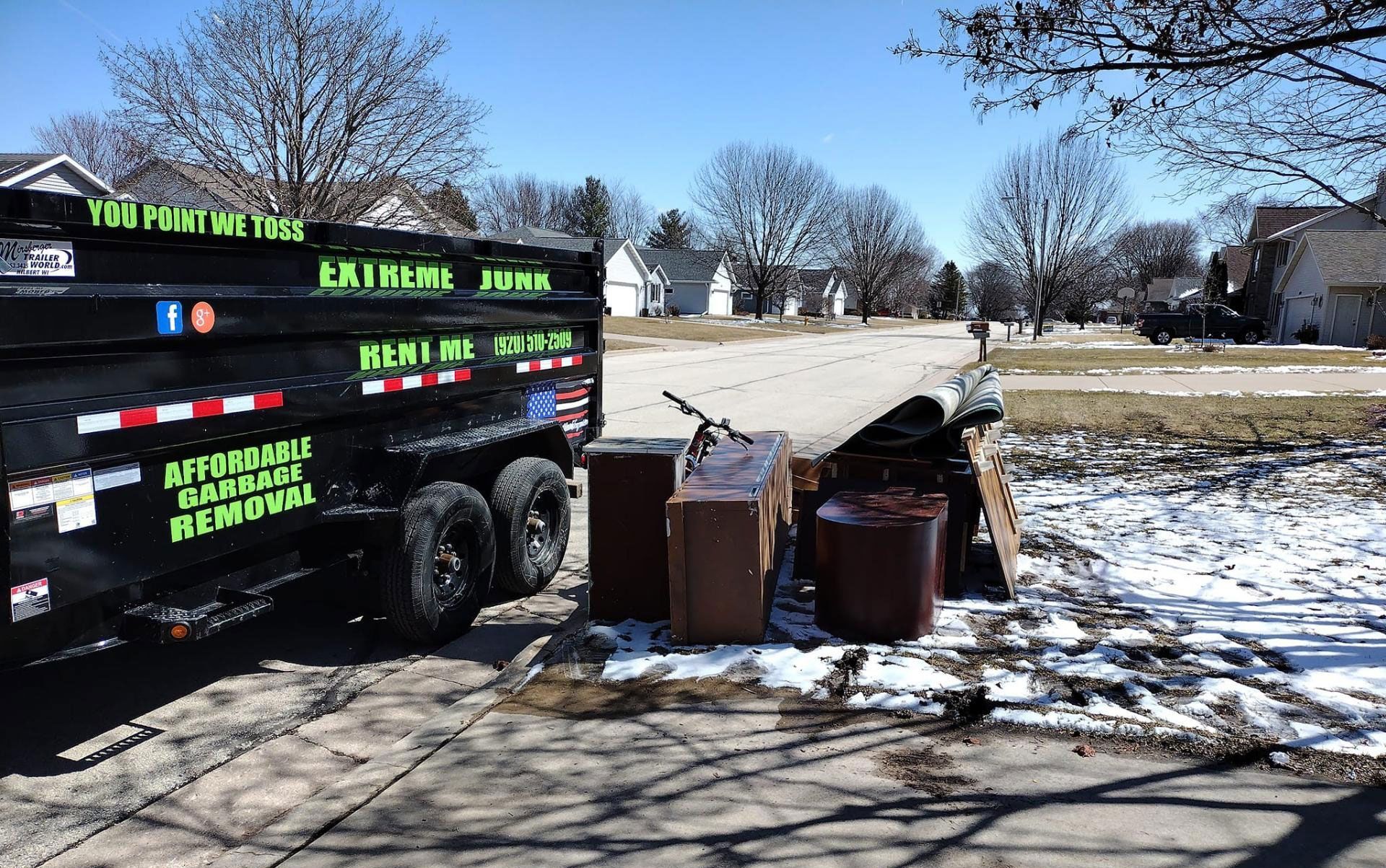 A black junk removal trailer parked on a suburban street next to a pile of old furniture and items on the side of the road.