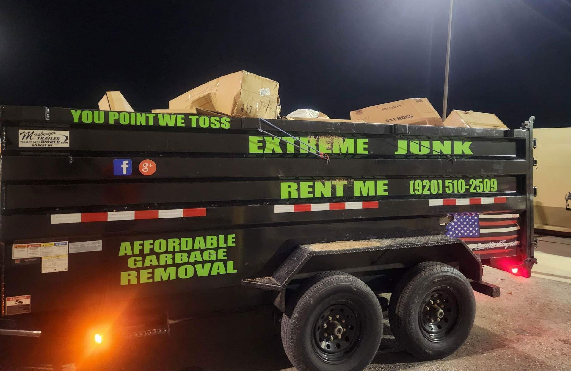 A black dump trailer for Extreme Junk, labeled with services and phone number, sits parked at night filled with cardboard.