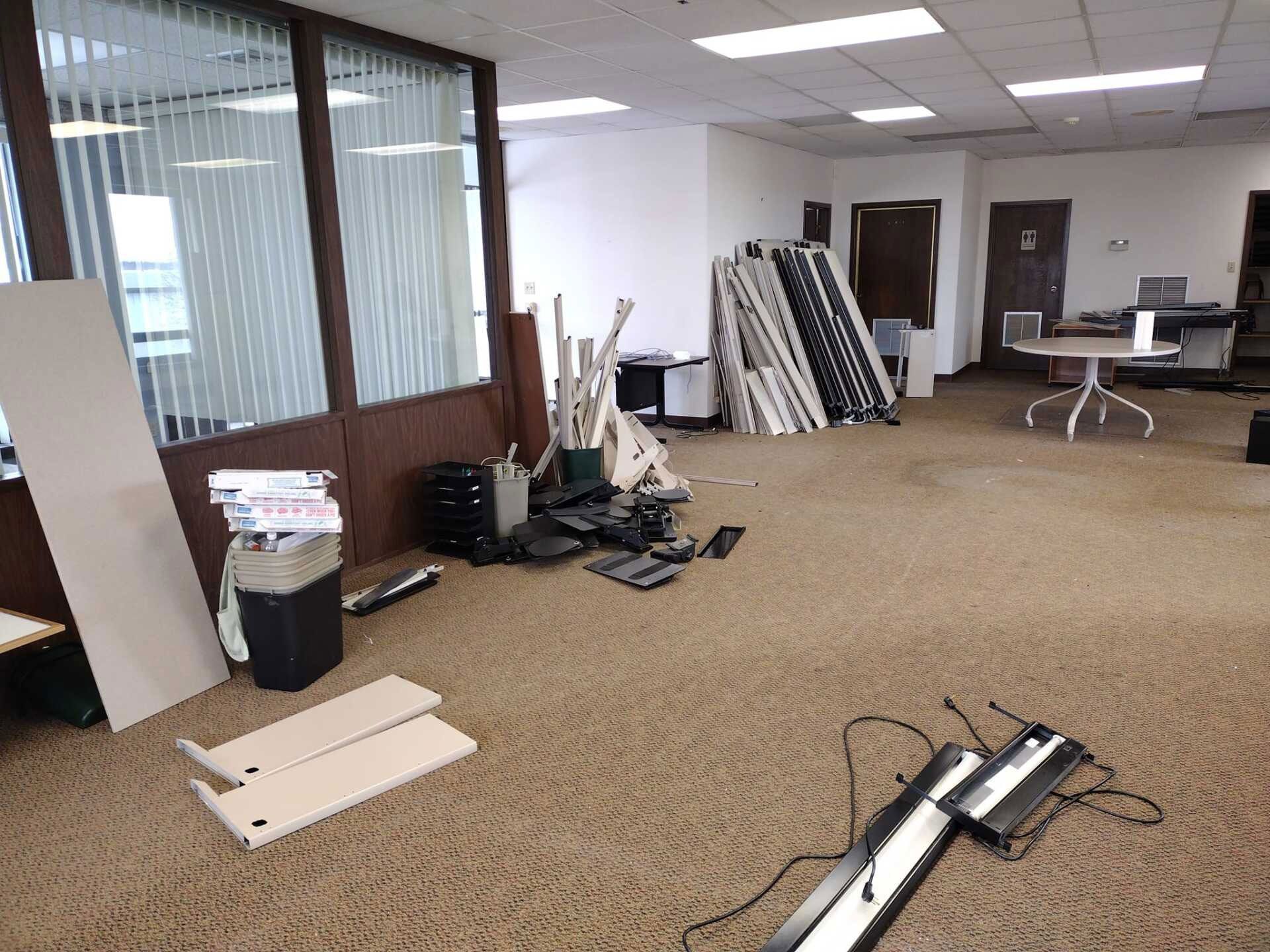An office under renovation with beige carpet, scattered furniture pieces, a stack of office chairs, and a round desk.