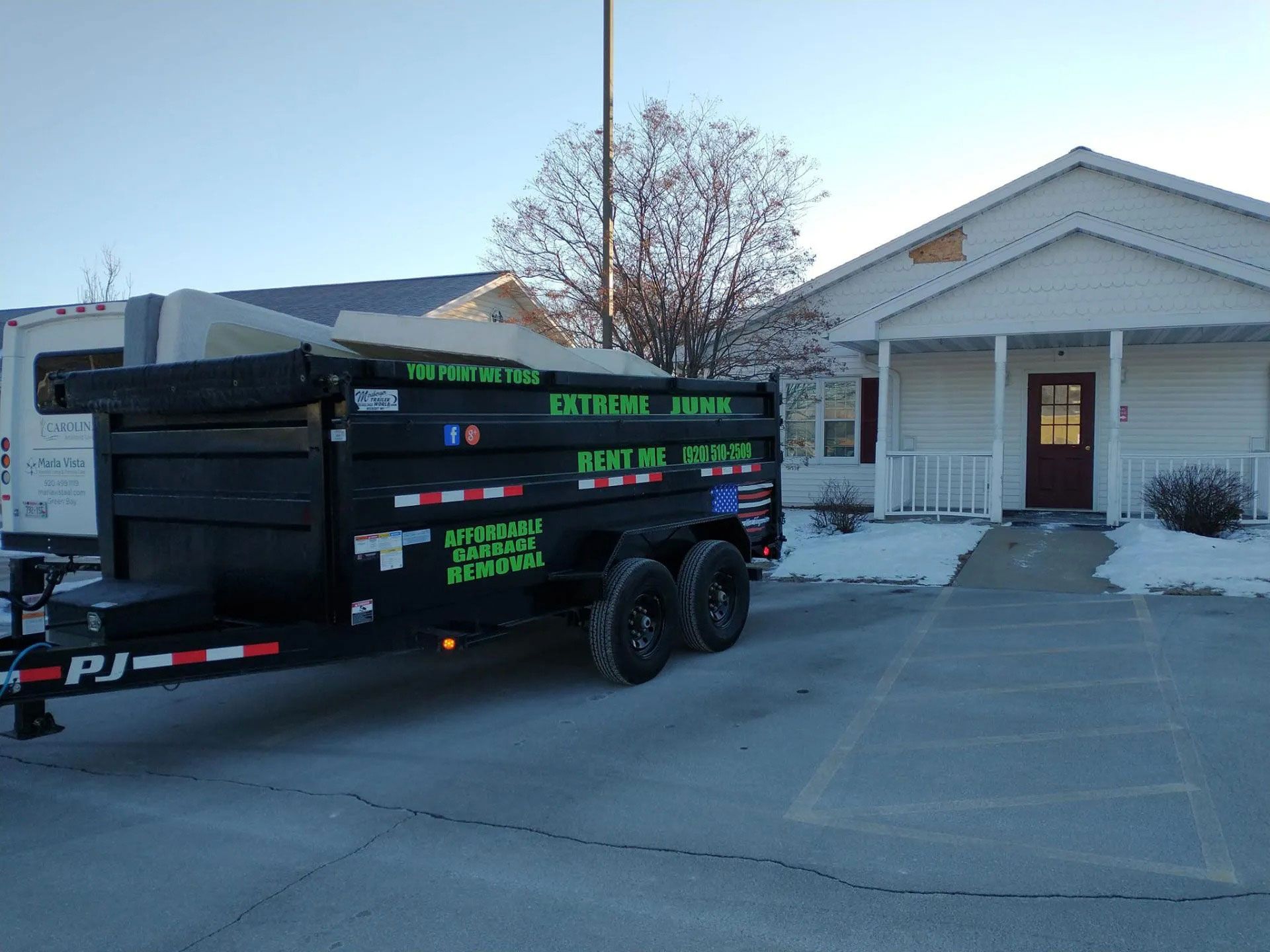 A black Extreme Junk trailer parked in front of a white house with a porch on a snowy day.
