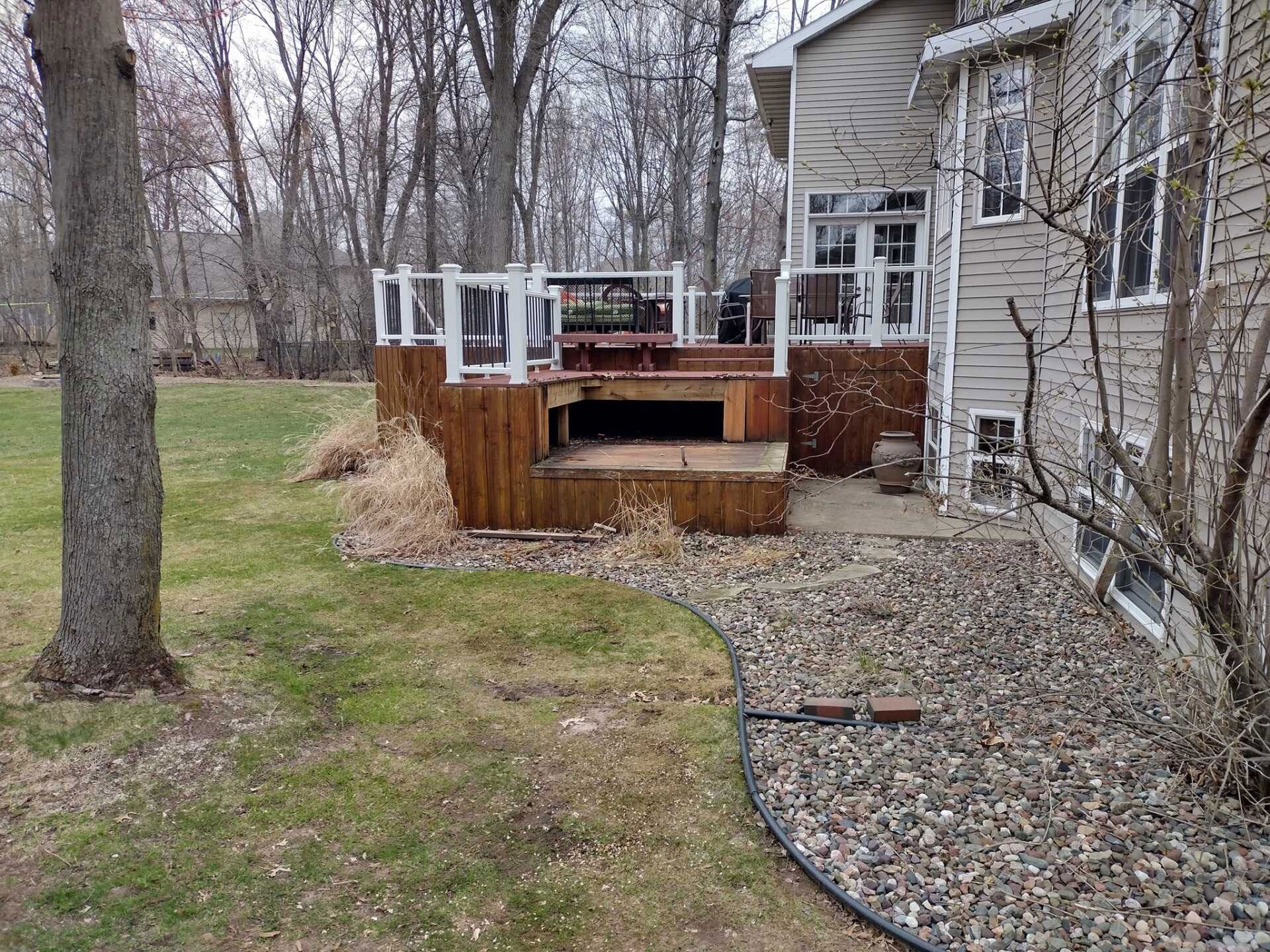 A two-level wooden deck attached to the side of a tan house, overlooking a grassy yard with trees and a stone border.