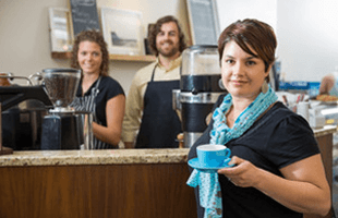 Portrait of young female customer holding coffee cup with workers in background at cafe counter