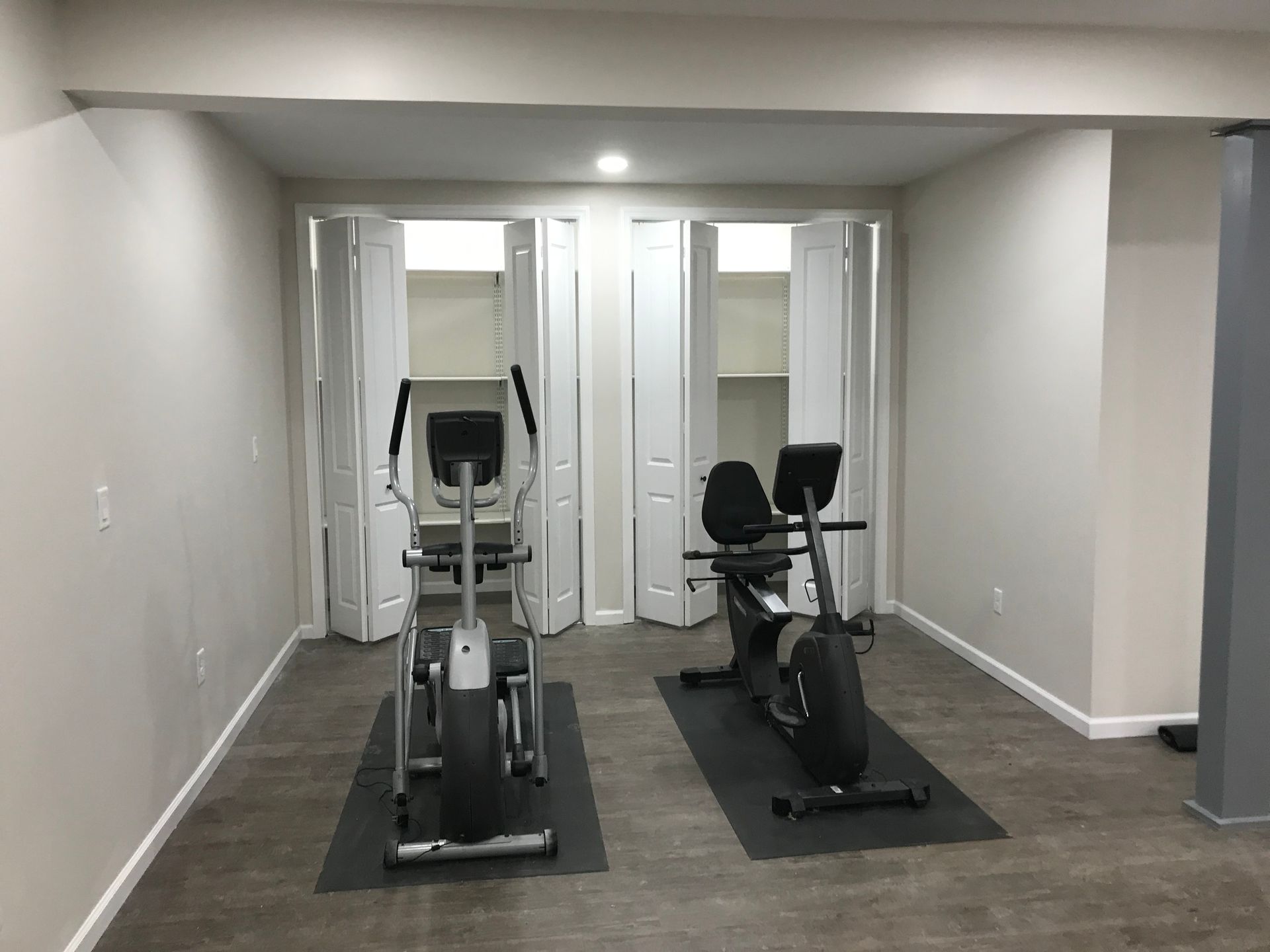 Home gym with elliptical machine and recumbent bike on mats. White bi-fold doors, neutral walls.