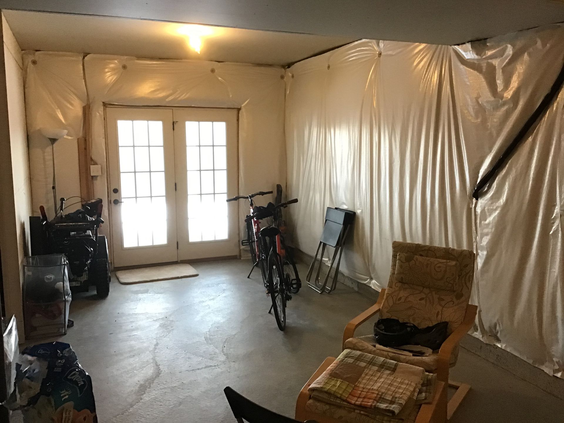 Basement room with French doors, bicycle, chair, plastic sheeting on walls, concrete floor.