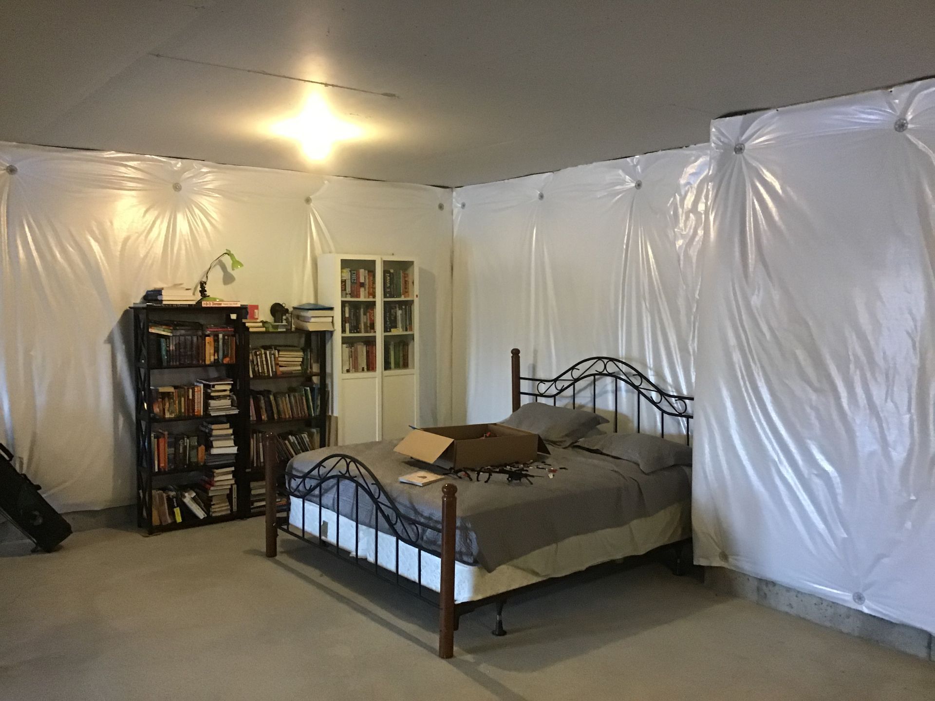 A makeshift bedroom with plastic sheeting walls, a bed, and bookshelves against the wall.