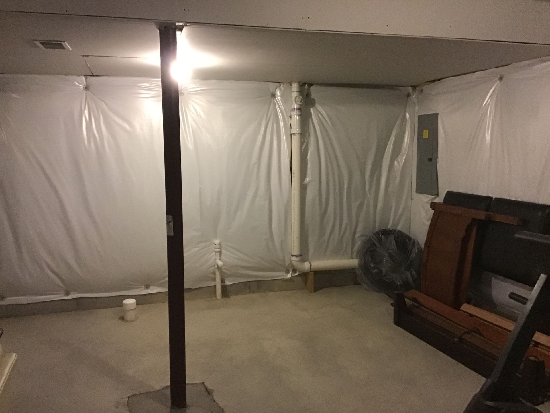 Basement interior with plastic sheeting on walls, support beam, electrical box, and discarded furniture.