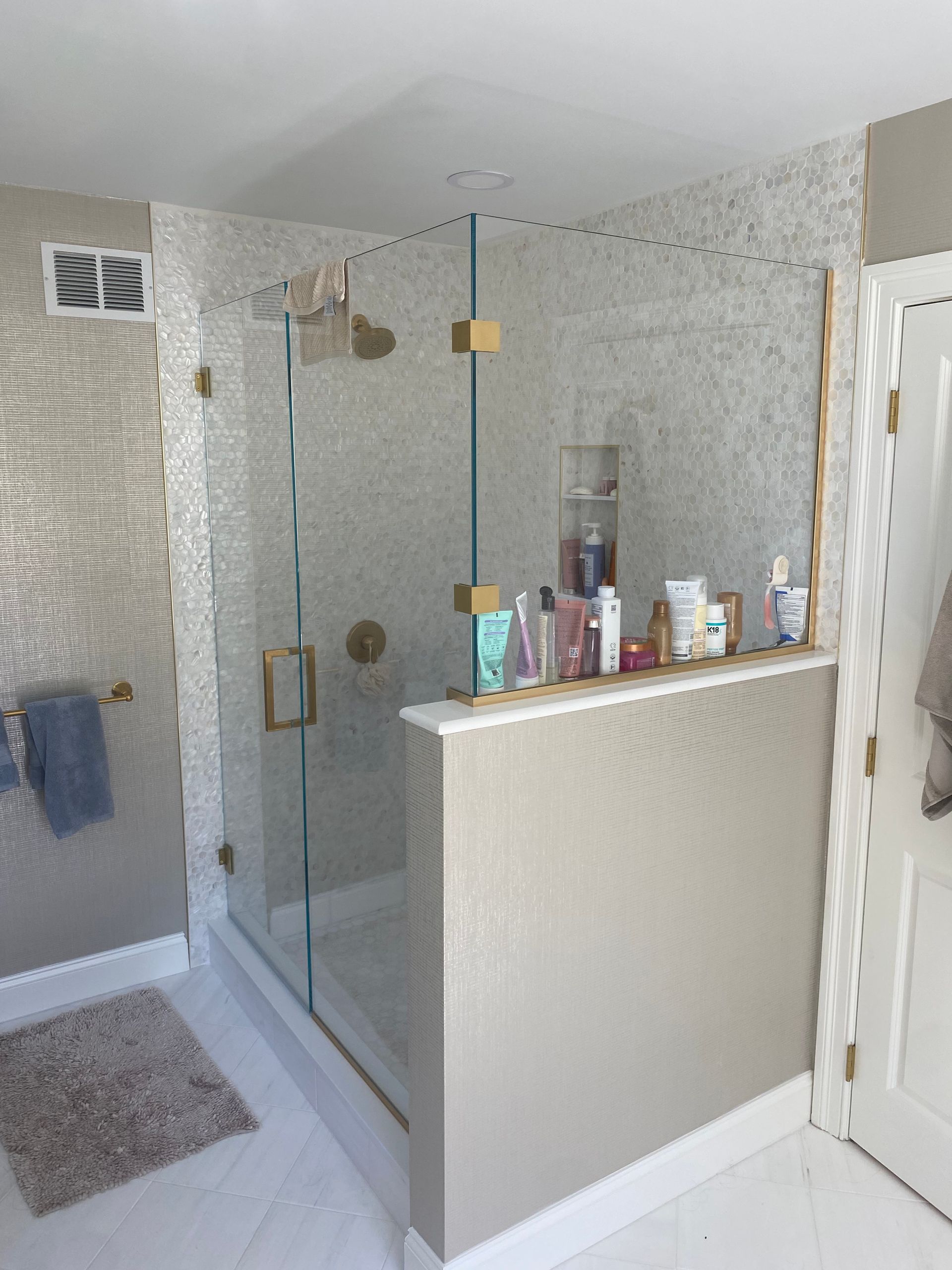 Shower with glass door, gold fixtures, beige tile walls, and a shelf with toiletries.