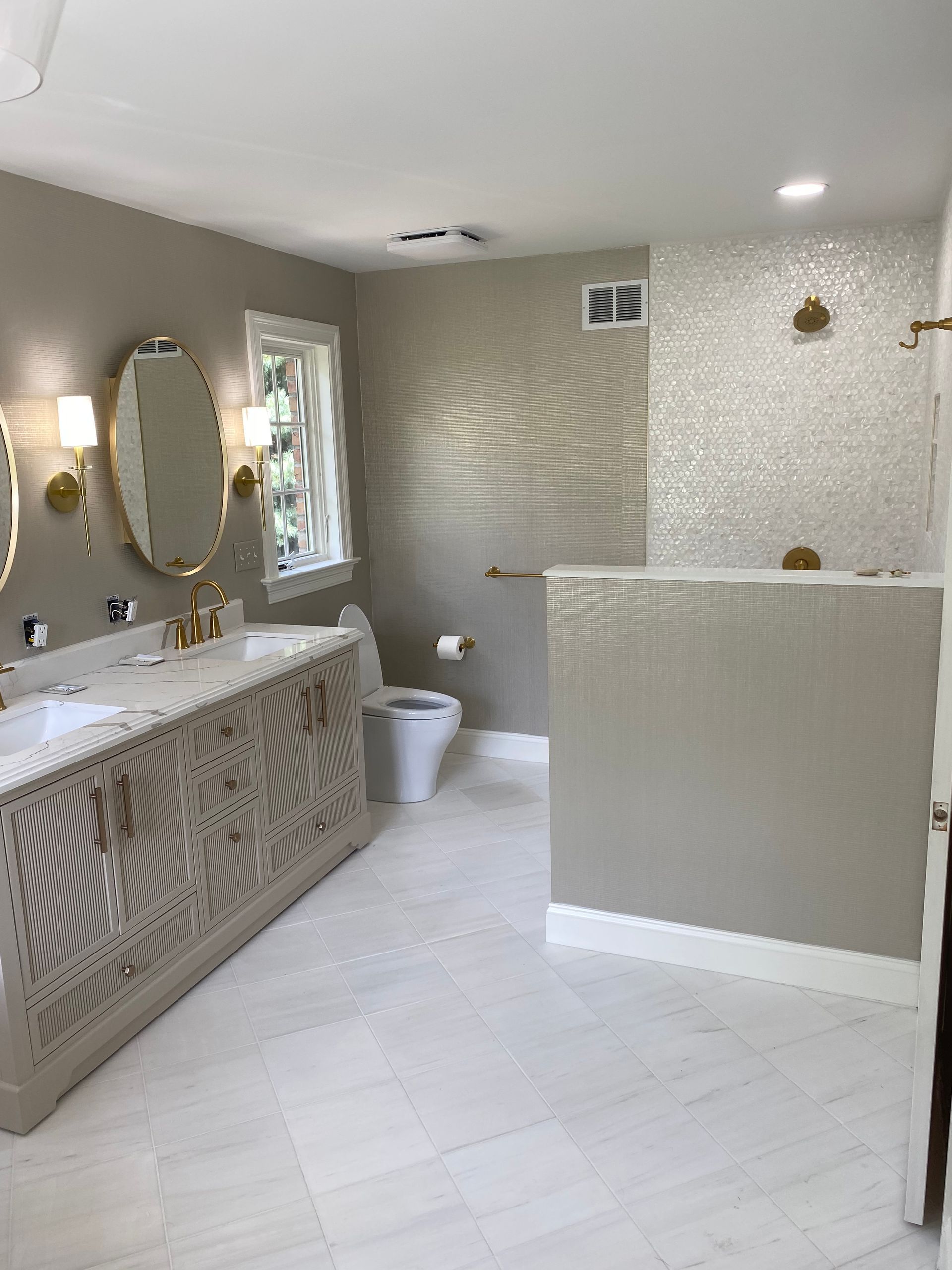 Bathroom with double vanity, toilet, and shower. Tan walls, white floor, gold fixtures.
