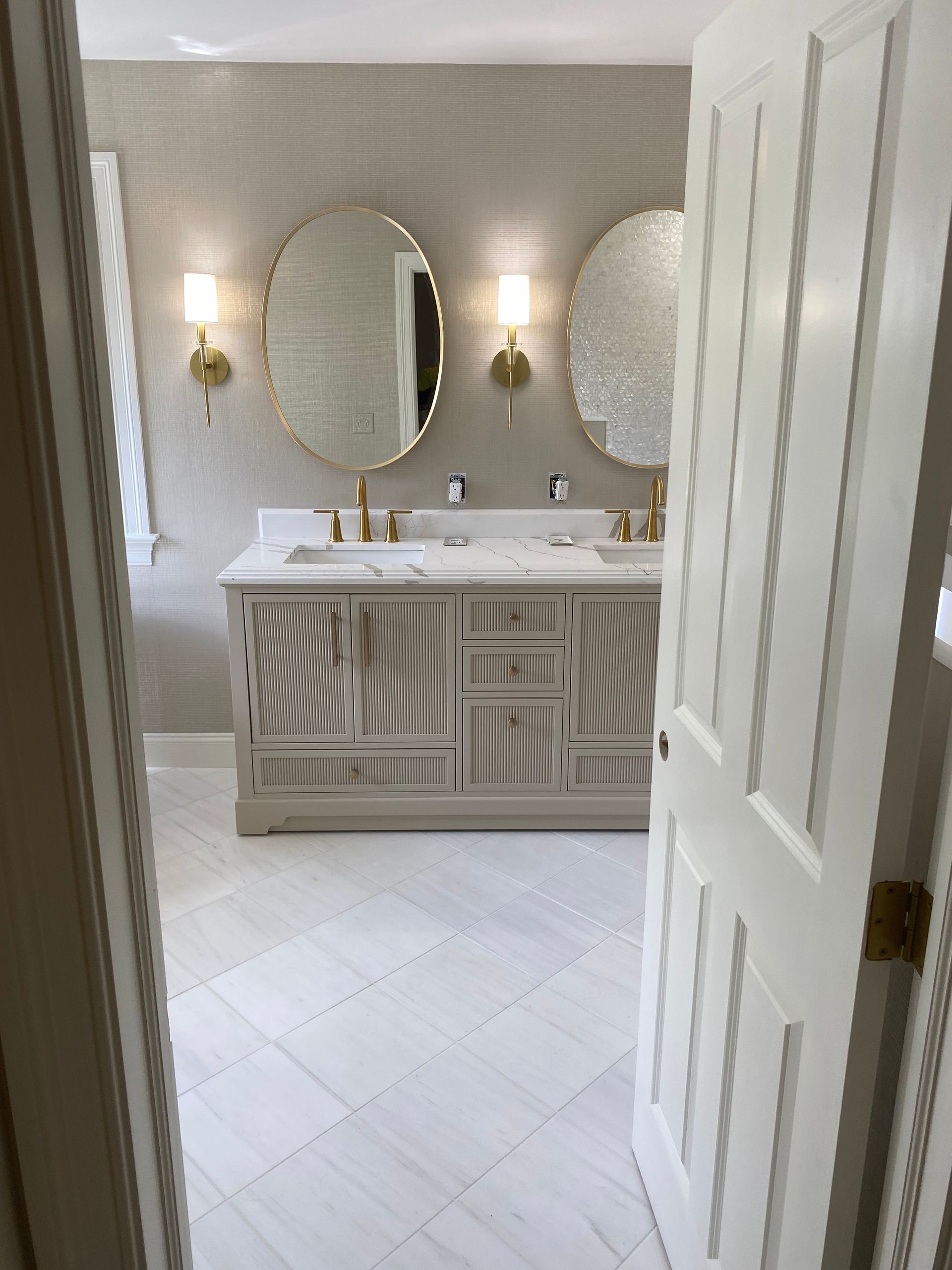 Bathroom with double sink vanity, oval mirrors, gold fixtures, and textured tile walls.