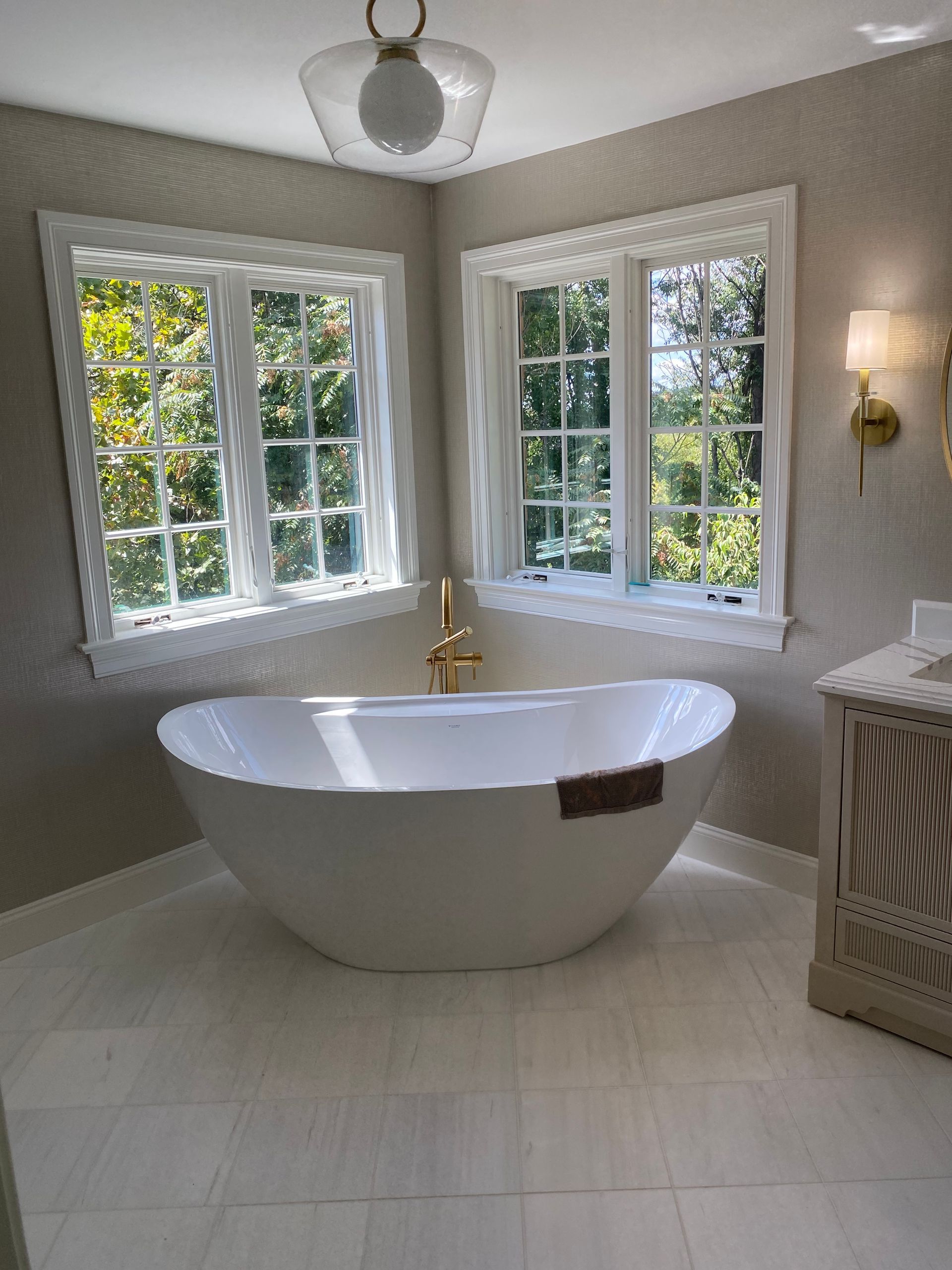 Bathroom with white bathtub under two windows overlooking trees. Gold fixtures, light beige walls, and white floor tiles.