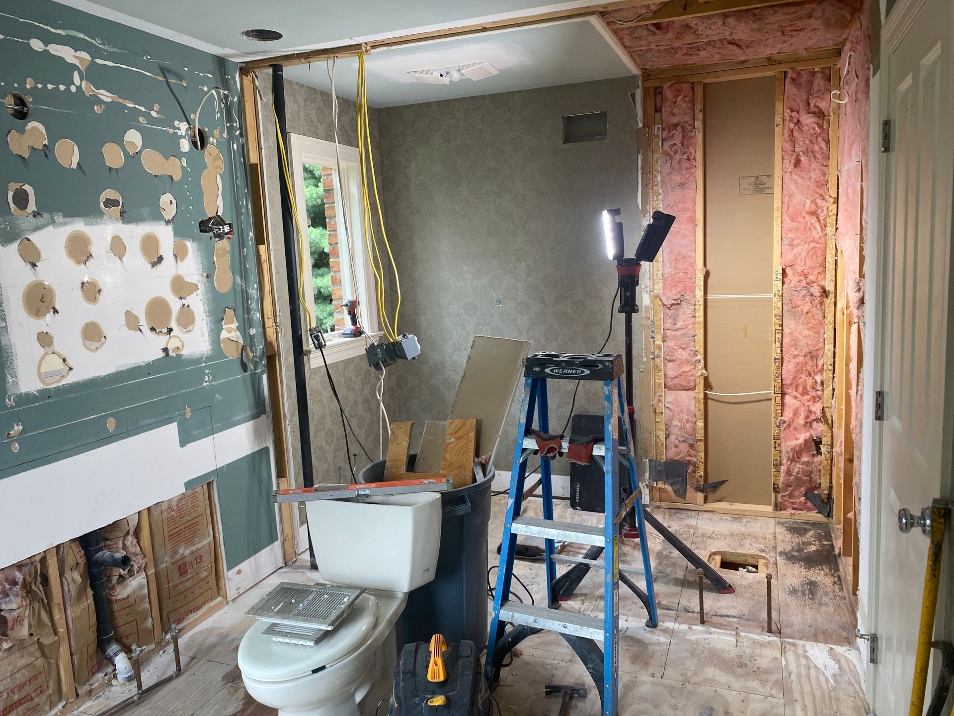 Bathroom renovation in progress, walls exposed, toilet installed, ladder and work lights visible.
