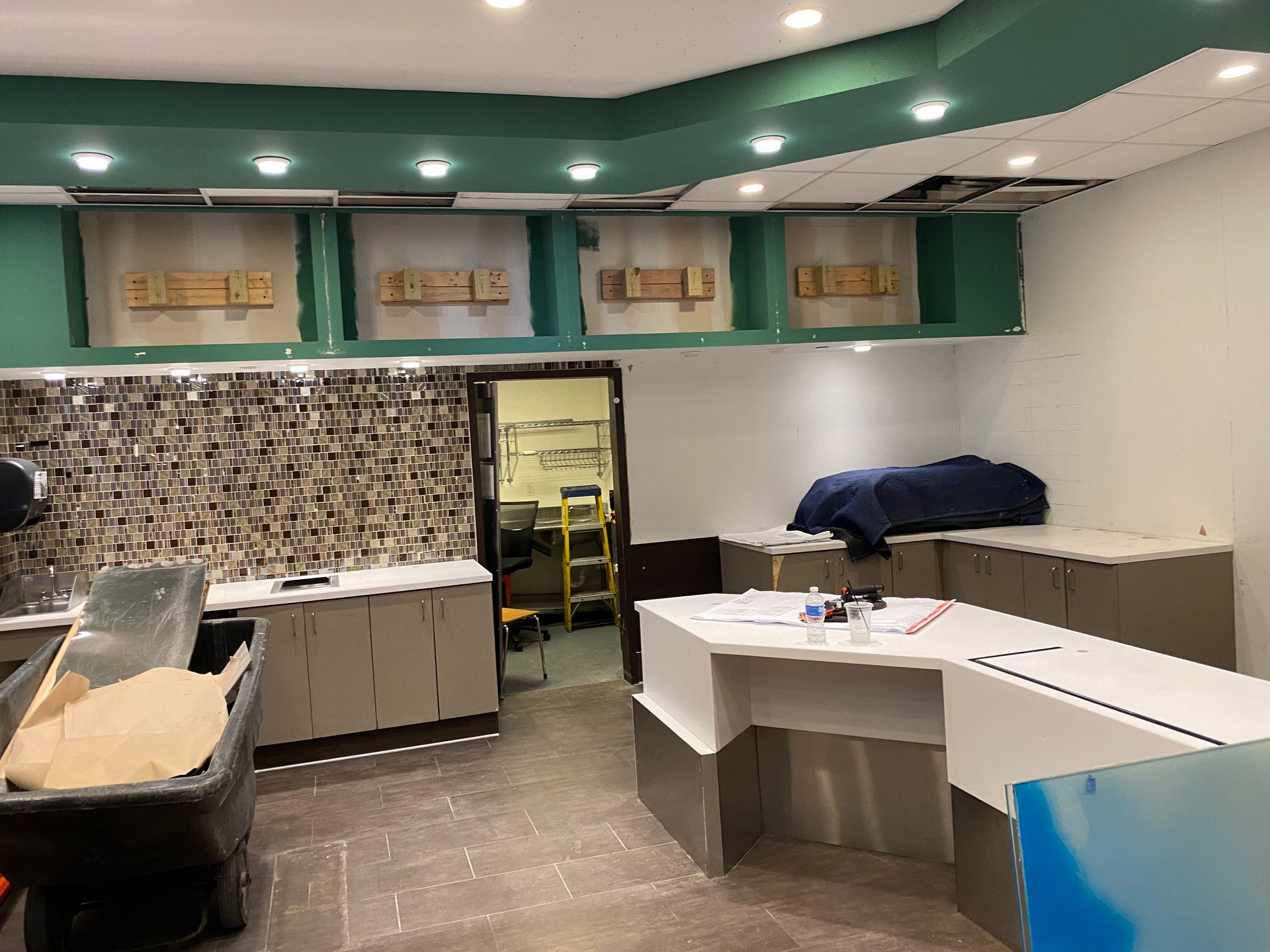Interior view of a commercial space under renovation. Green and white walls, cabinetry, and a service counter are visible.