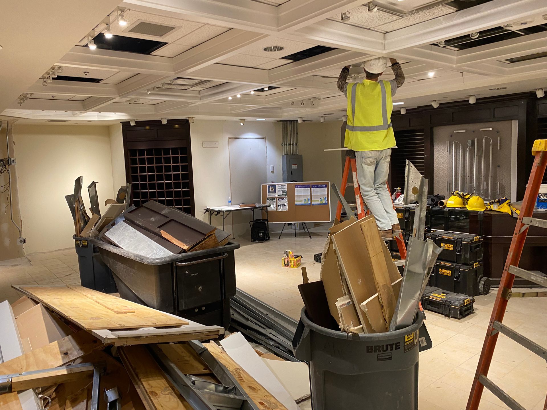 Construction worker on ladder working on a ceiling in a room under renovation, tools and debris visible.