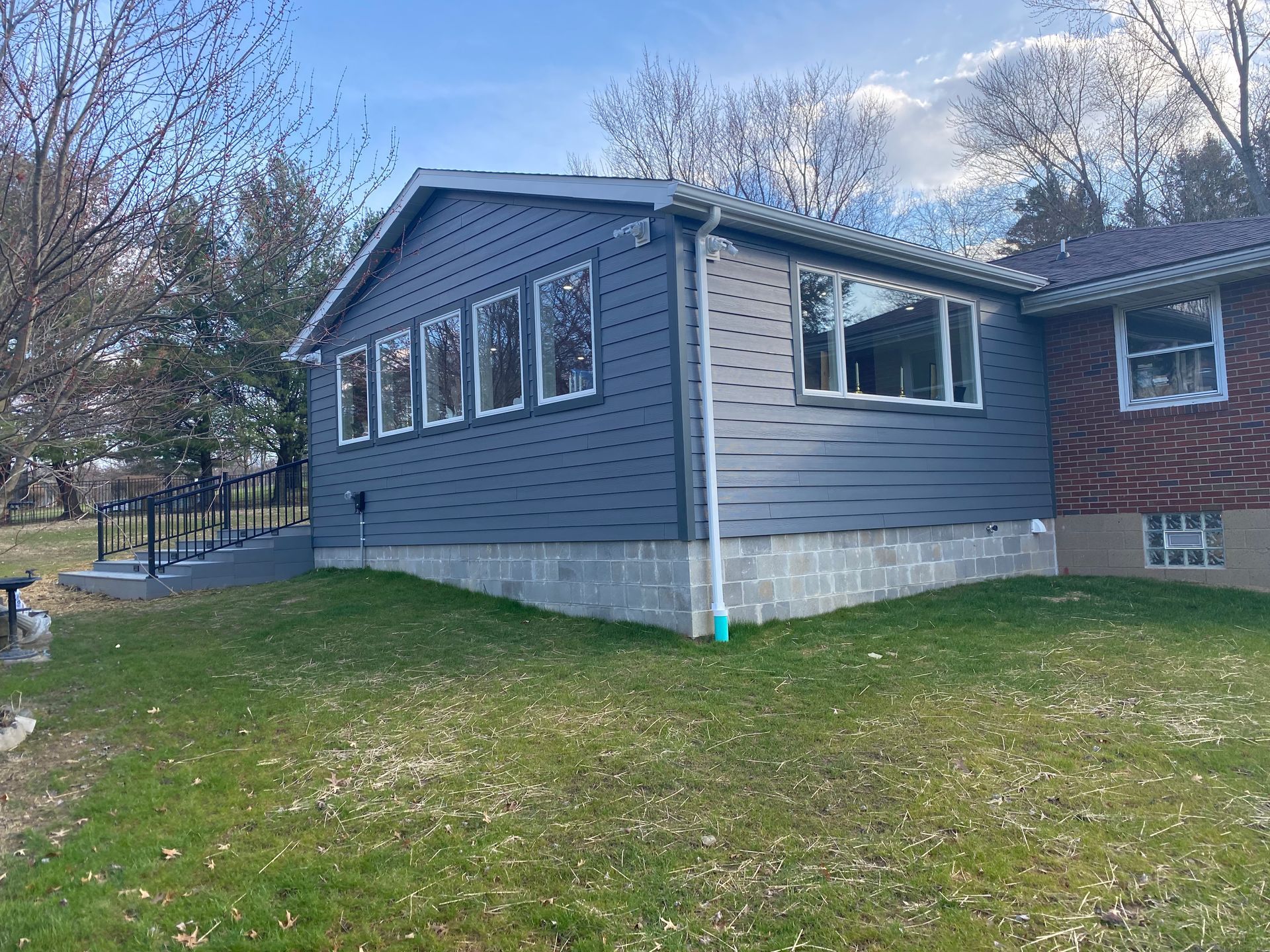 A house with gray siding addition and brick section, on a grassy lawn.