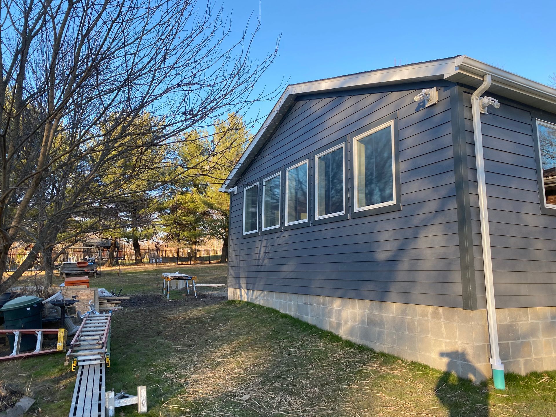 Dark gray house with several windows, white trim and gutter, on a concrete foundation; outdoors, sunny.