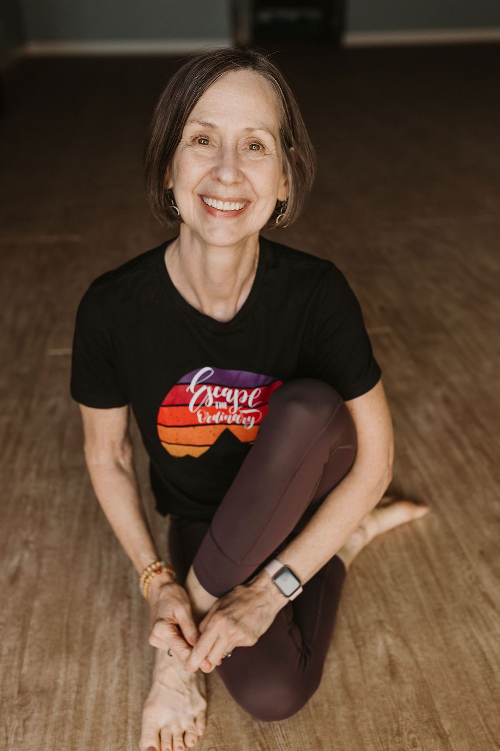 Smiling woman seated cross-legged on a wooden floor, wearing a black T-shirt and maroon leggings.